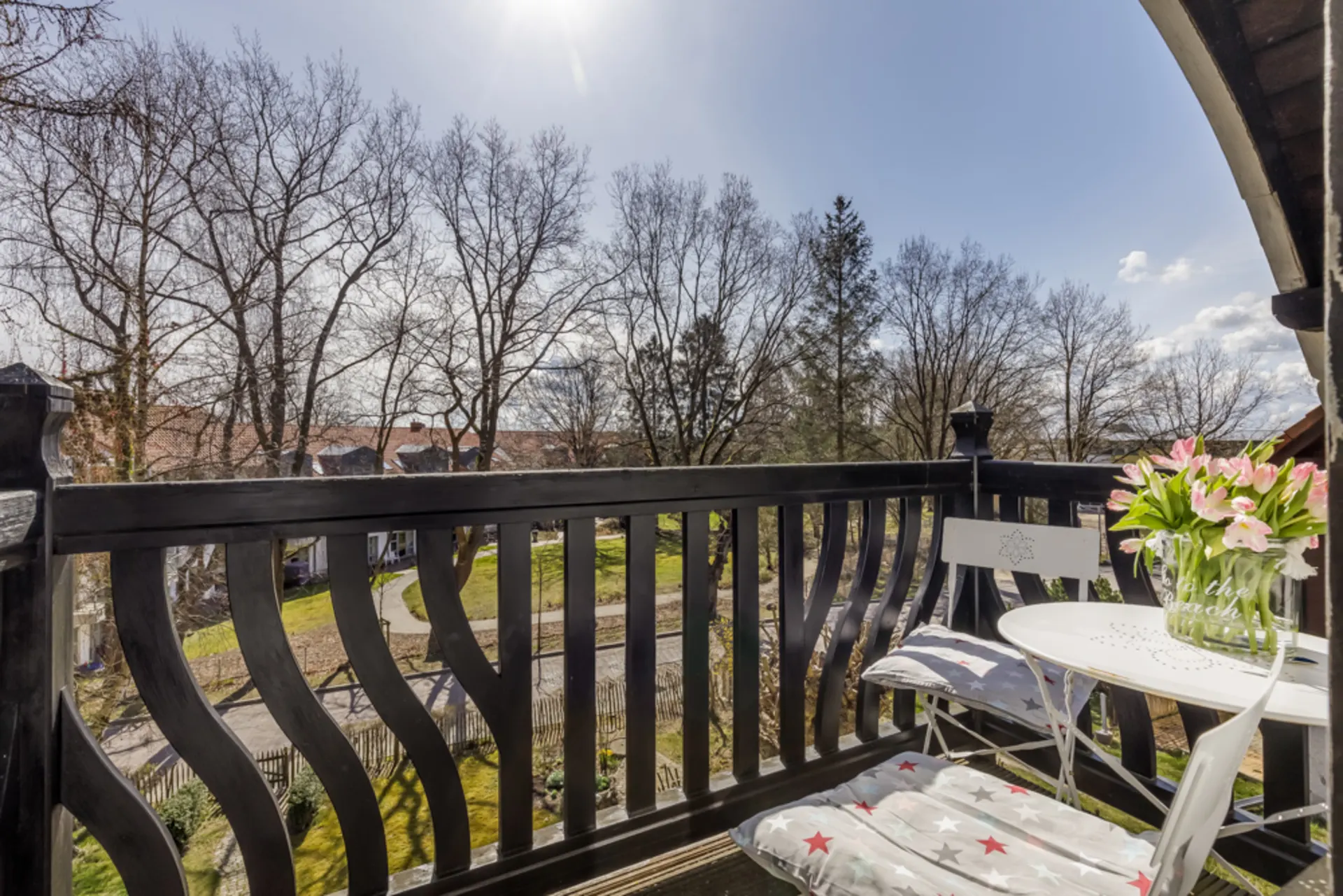 Kleiner Balkon mit Tisch, zwei Stühlen und Blumen, Blick auf Garten und Bäume im Sonnenschein.