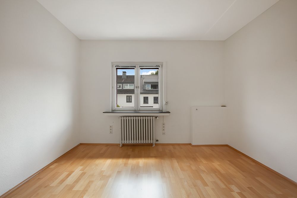 Leeres Zimmer mit hellen Wänden, Holzboden und einem Fenster mit Blick nach draußen.