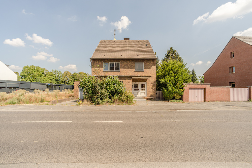 Zweistöckiges Wohnhaus mit Satteldach und zwei Eingängen, umgeben von Büschen, an einer Straße gelegen.