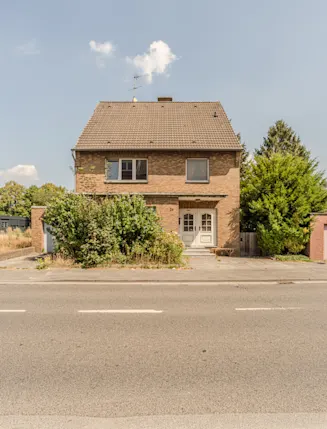 Zweistöckiges Wohnhaus mit Satteldach und zwei Eingängen, umgeben von Büschen, an einer Straße gelegen.