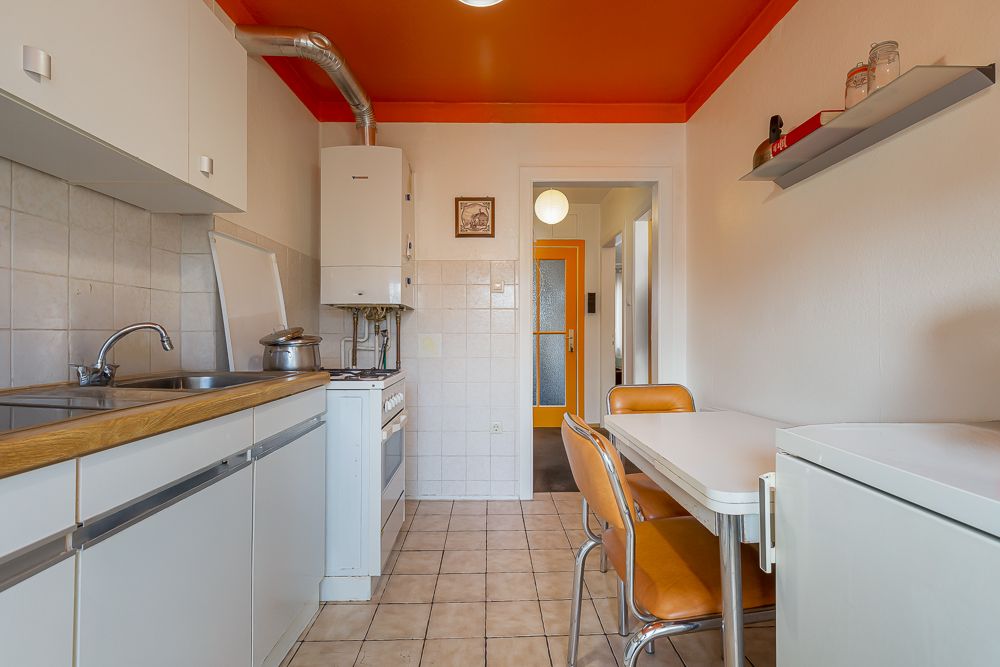 Helle Küche mit weißen Schränken, Esstisch, orangener Decke und Fliesenboden.