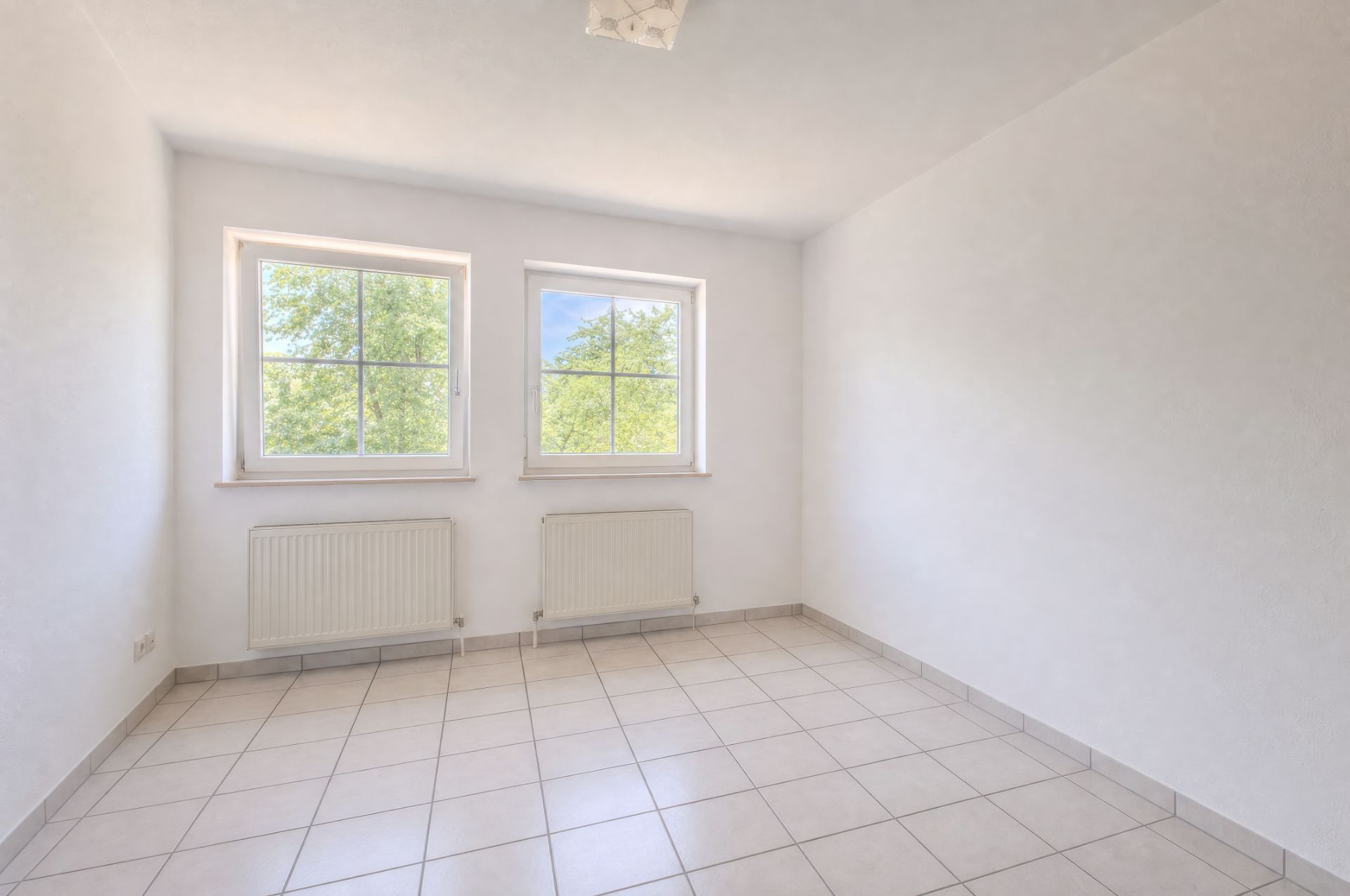Helles, leeres Zimmer mit zwei Fenstern, weißen Wänden und Fliesenboden.