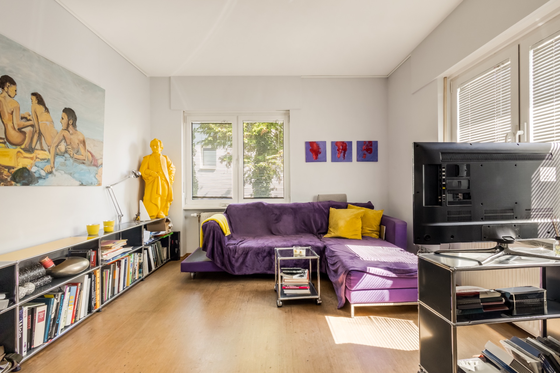 Wohnzimmer mit lila Sofa, gelben Kissen, Bücherregal und großem TV, Kunst an den Wänden.