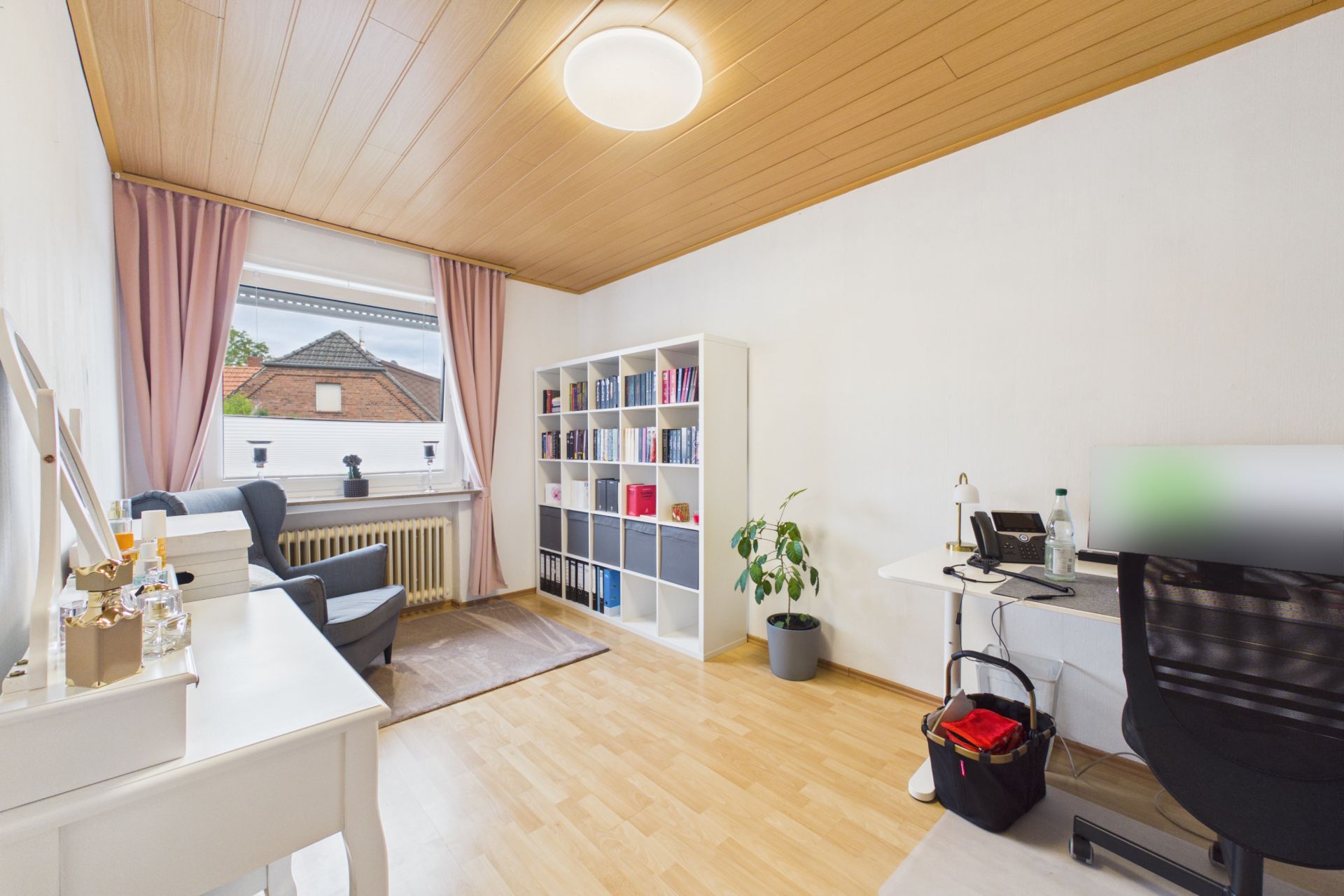Helles Zimmer mit Schreibtisch, Regal, Sessel, großem Fenster und Holzdecke.