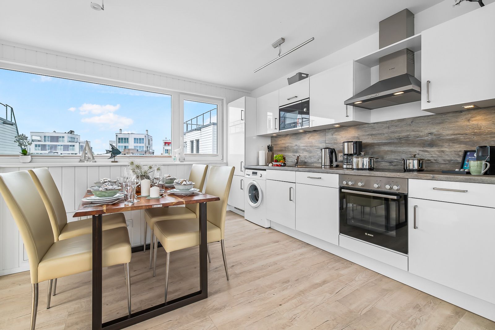 Moderne Küche mit Esstisch und vier Stühlen, große Fenster mit Blick auf Häuser.
