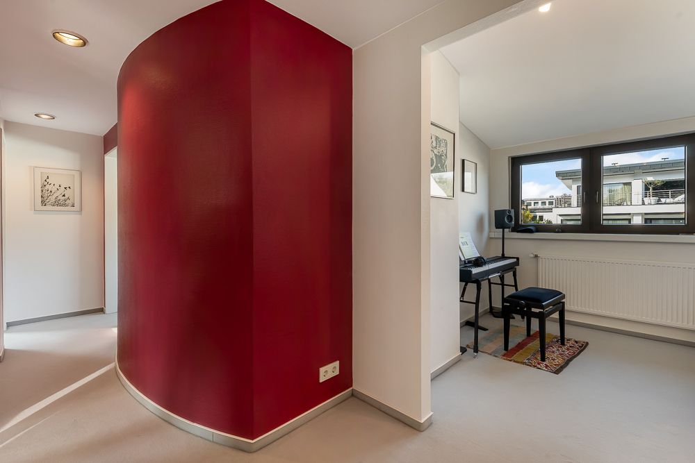 Flur mit roter Rundwand und Blick in ein Zimmer mit Piano und großem Fenster.