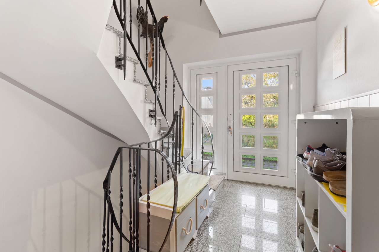 Heller Flur mit Treppe, Schuhregal und Sitzbank, große Glastür im Hintergrund.