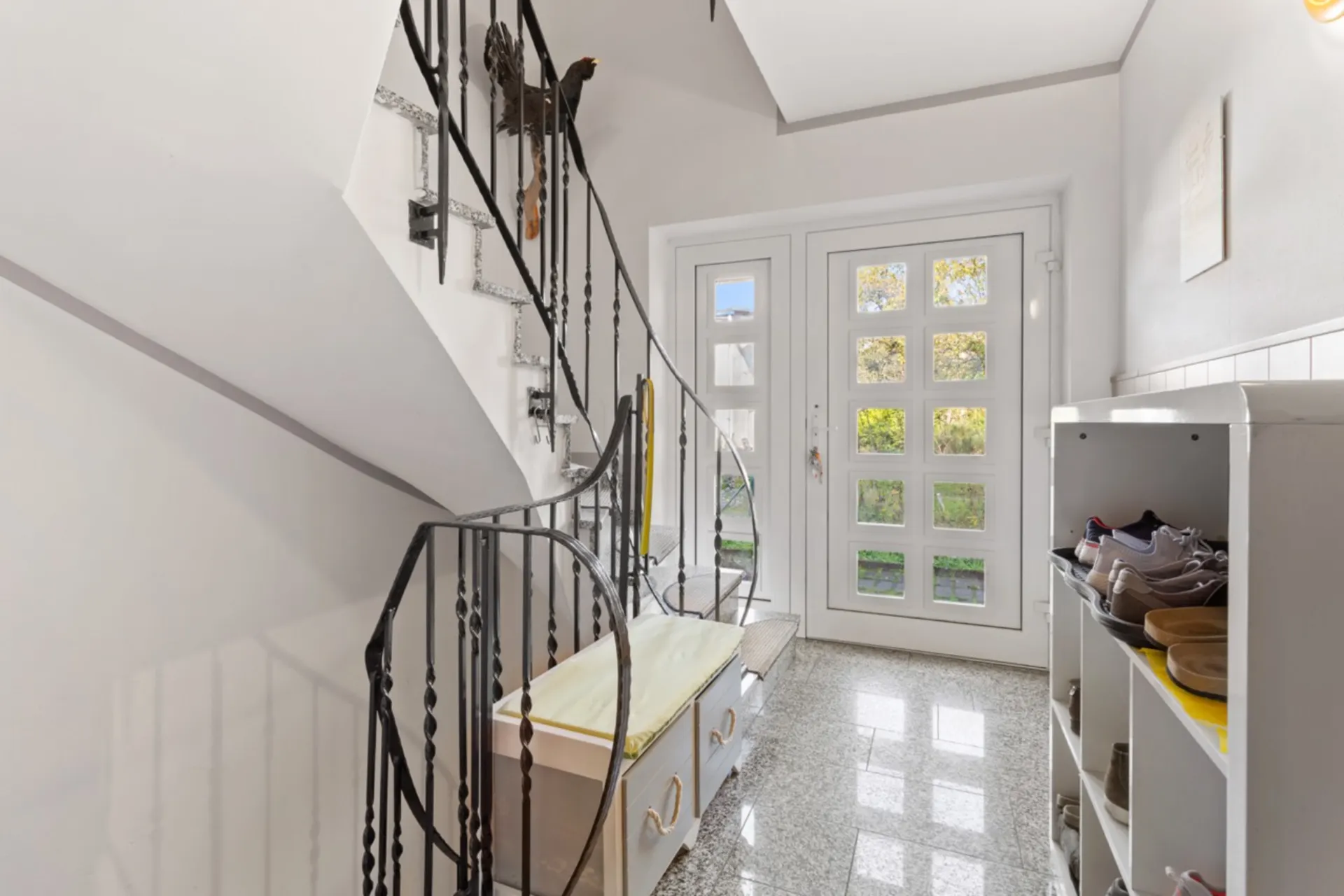 Heller Flur mit Treppe, Schuhregal und Sitzbank, große Glastür im Hintergrund.