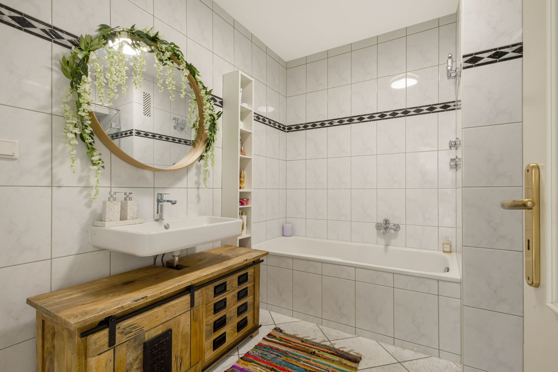 Helles Badezimmer mit Badewanne, Holzwaschtisch, rundem Spiegel und gemustertem Teppich.