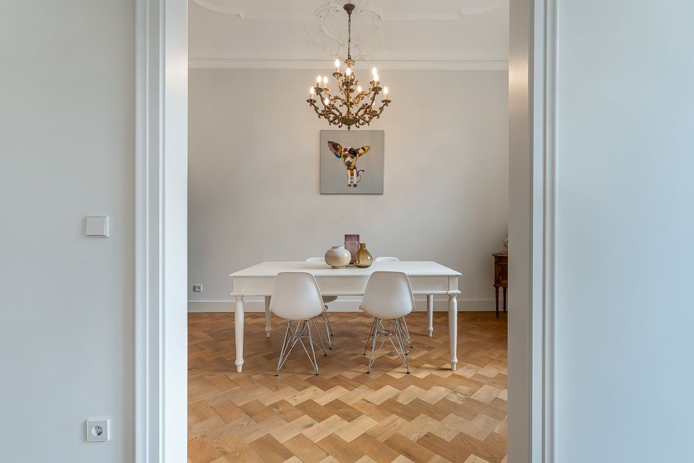 Esszimmer mit weißem Tisch, vier weißen Stühlen und Kronleuchter auf Parkettboden.