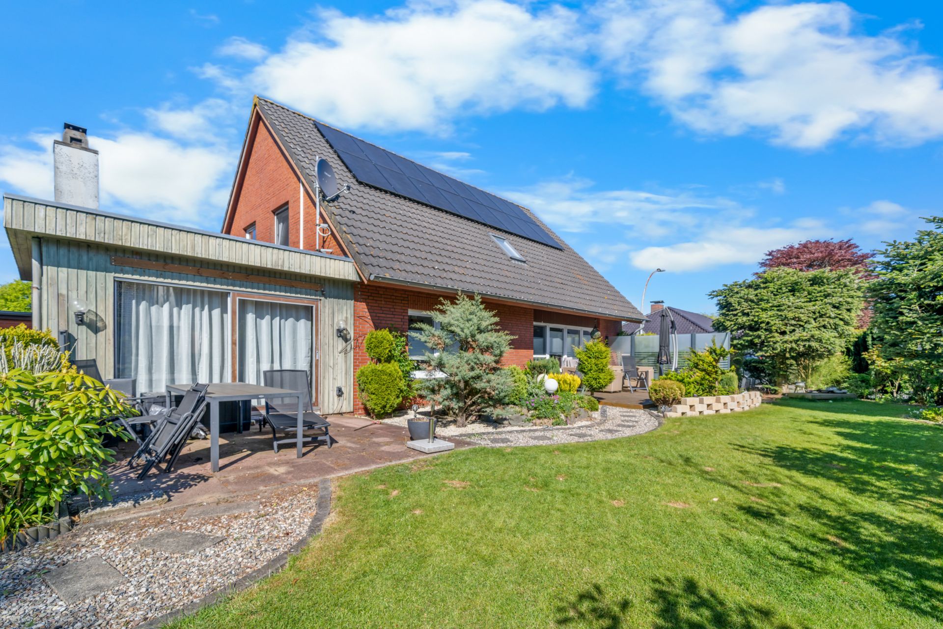 Haus mit Solarpanelen auf dem Dach, Terrasse mit Sitzgruppe und gepflegtem Garten bei sonnigem Wetter.