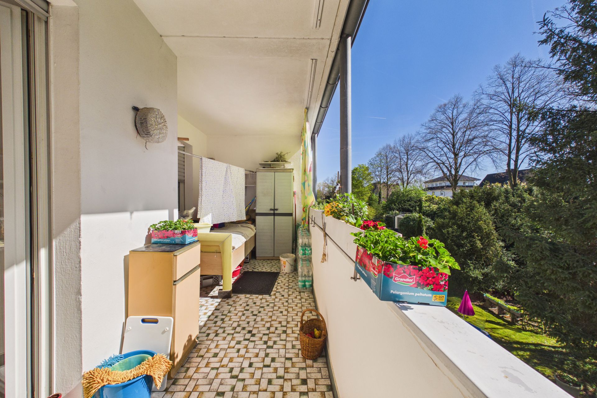 Langer Balkon mit Fliesenboden, Pflanzkästen, Schränken und Blick auf einen grünen Garten.