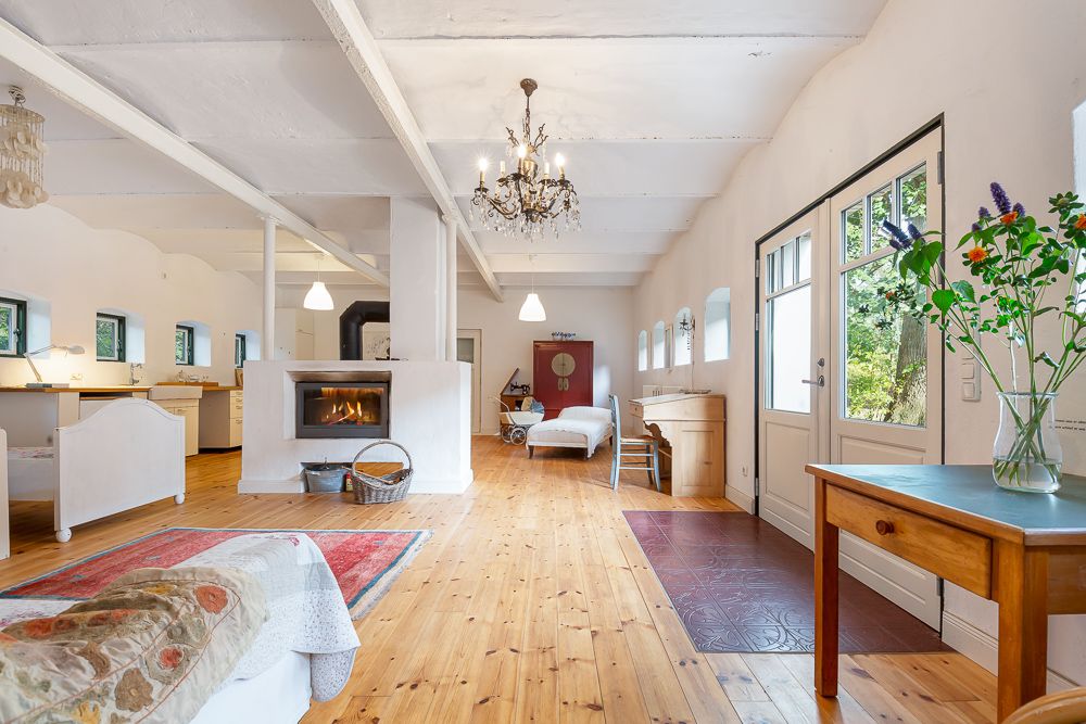 Heller Wohnraum mit Holzboden, Kamin, Esstisch, Betten und großem Fenster neben einer Tür.