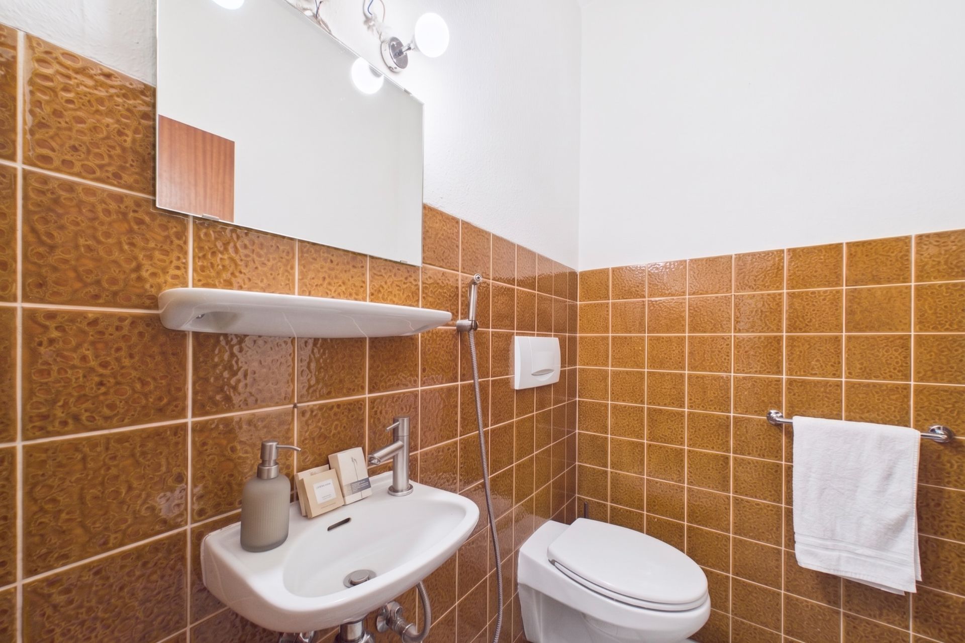 Kleines Badezimmer mit braunen Fliesen, Waschbecken, Spiegel, WC und Handtuchhalter.
