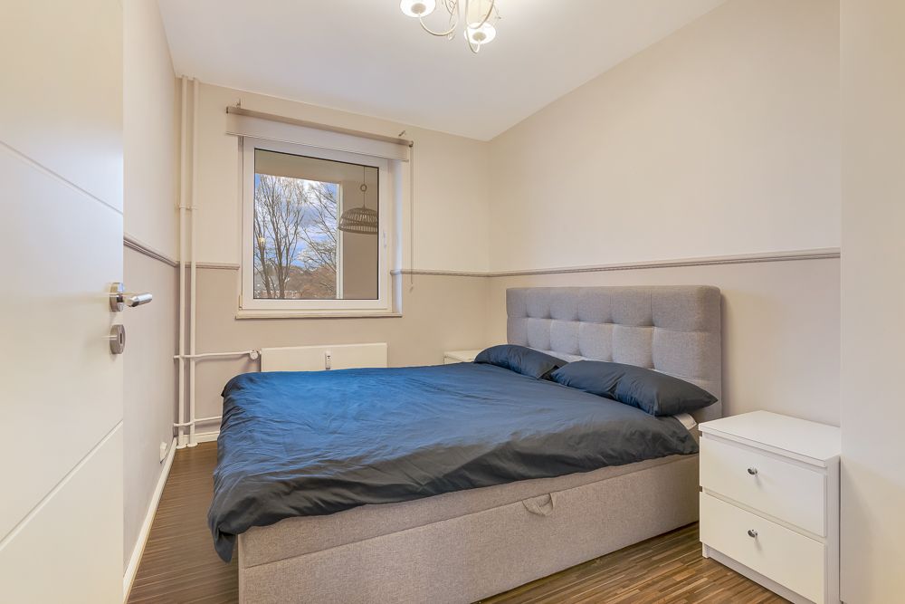Schlafzimmer mit grauem Bett, Nachttisch und Fenster mit Ausblick auf Bäume.