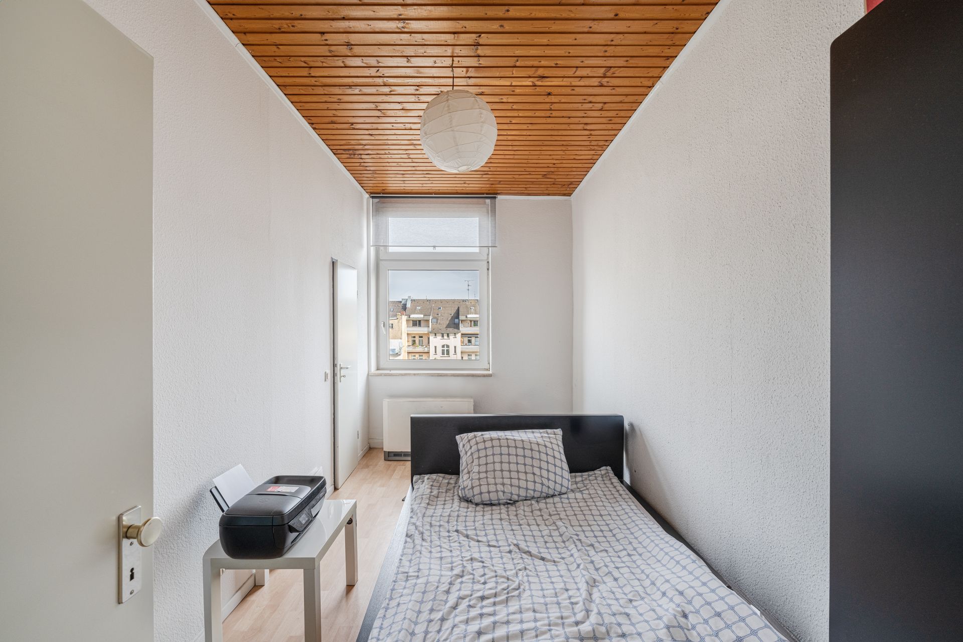 Schmales Schlafzimmer mit Einzelbett, kleinem Tisch und Fenster, Holzdecke und heller Wand.