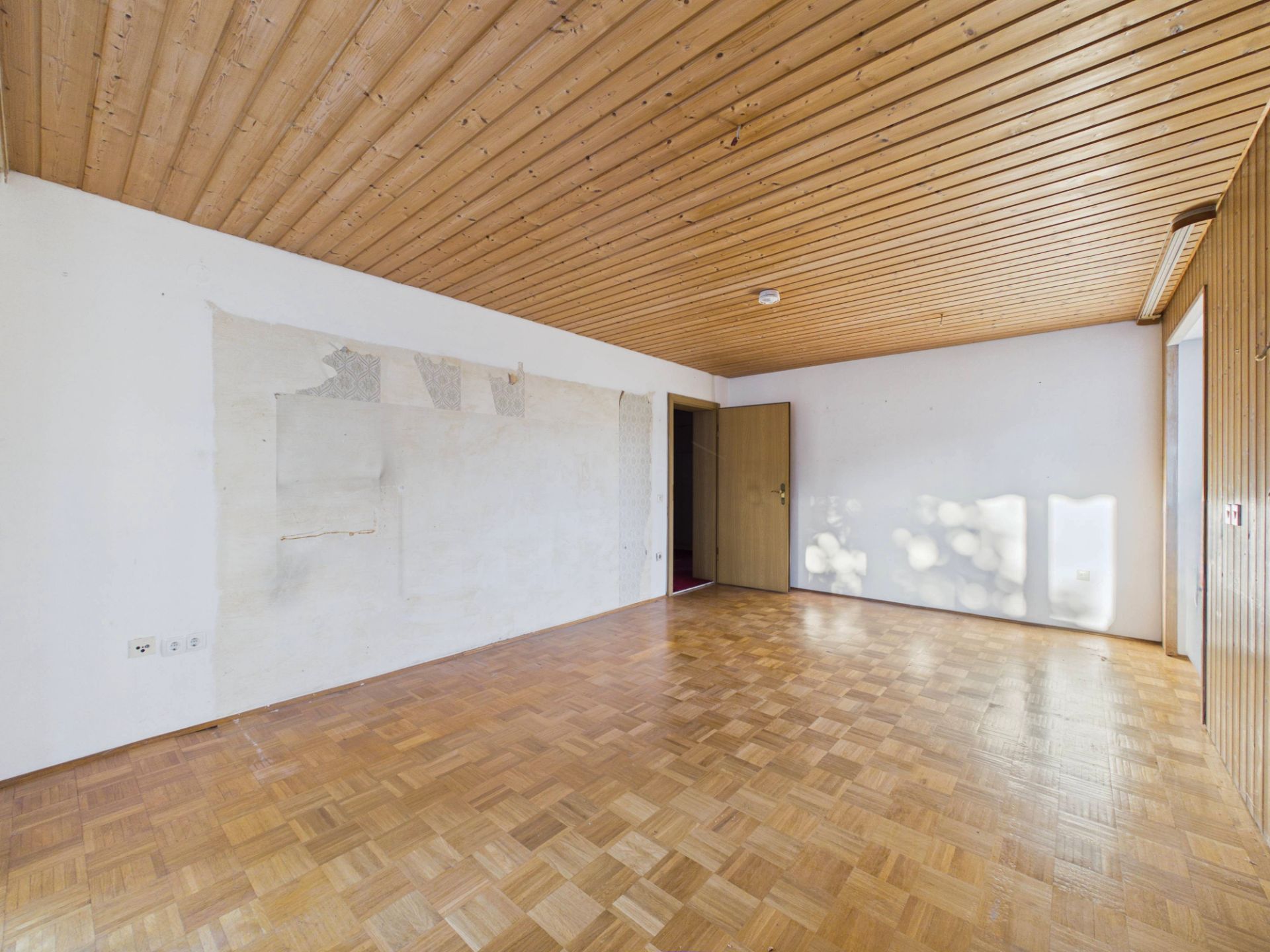 Leerstehendes Zimmer mit Parkettboden und Holzdecke, eine Wand teilweise beschädigt.