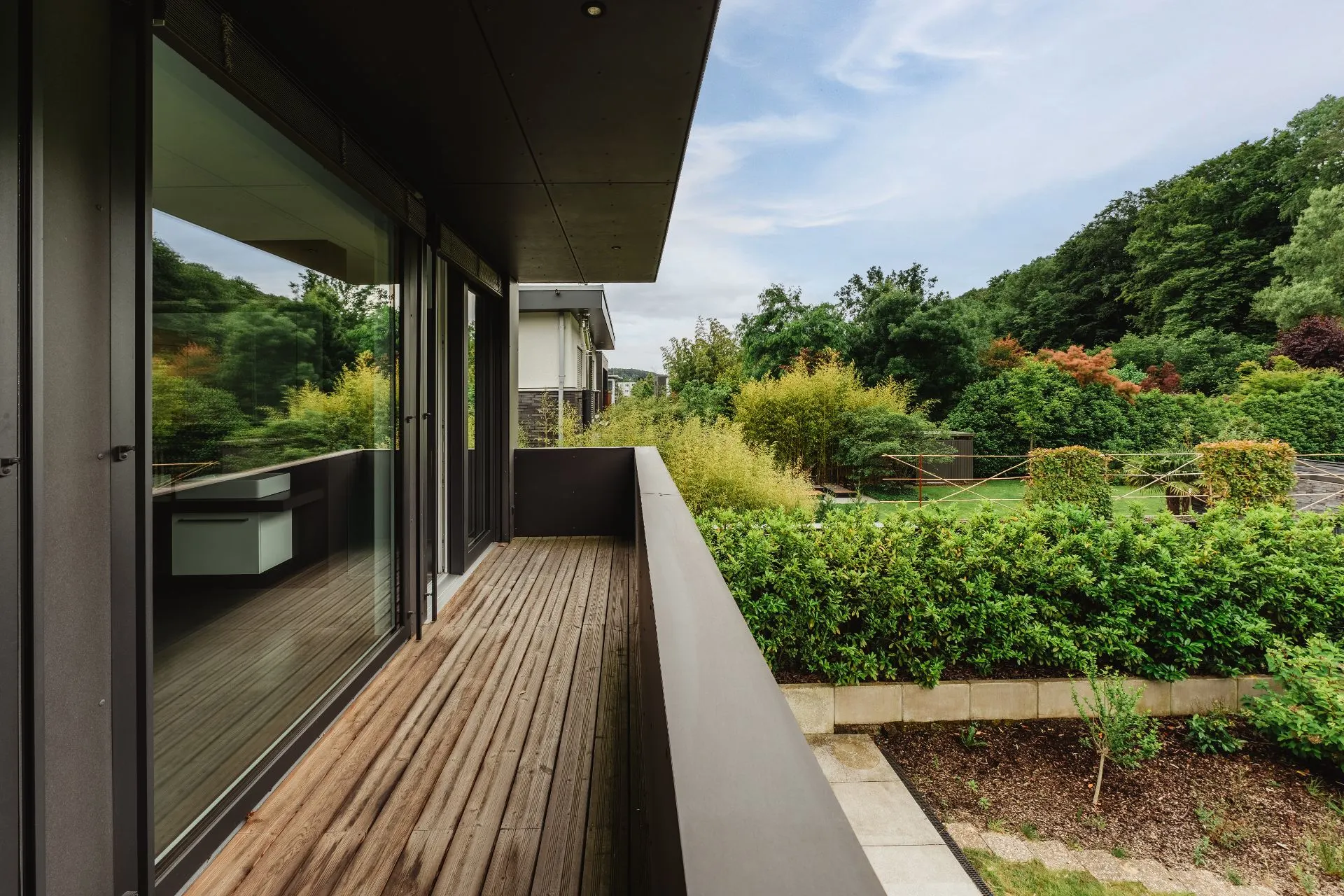 Moderner Balkon mit Holzfußboden, Blick auf Garten mit vielen grünen Büschen und Bäumen.