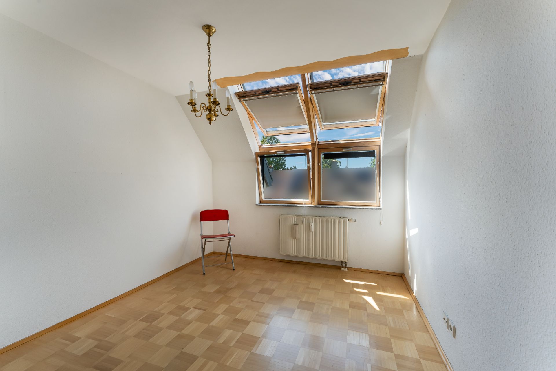 Leeres Zimmer mit Parkettboden, Dachfenster, Heizkörper und einem Stuhl mit roter Rückenlehne.
