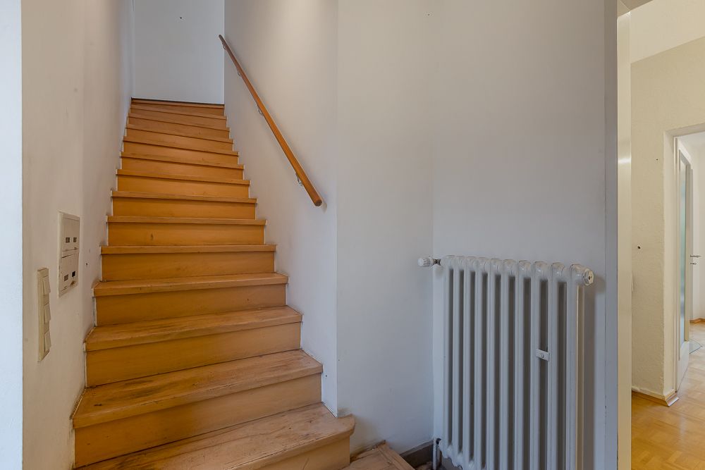 Holztreppe mit Handlauf neben einem Heizkörper in einem Flur mit weißen Wänden.