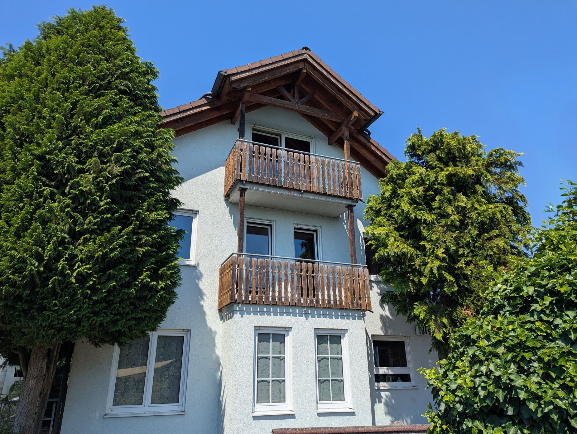 Haus mit zwei Holzbalkonen, weißen Fenstern und umgeben von Bäumen bei blauem Himmel.