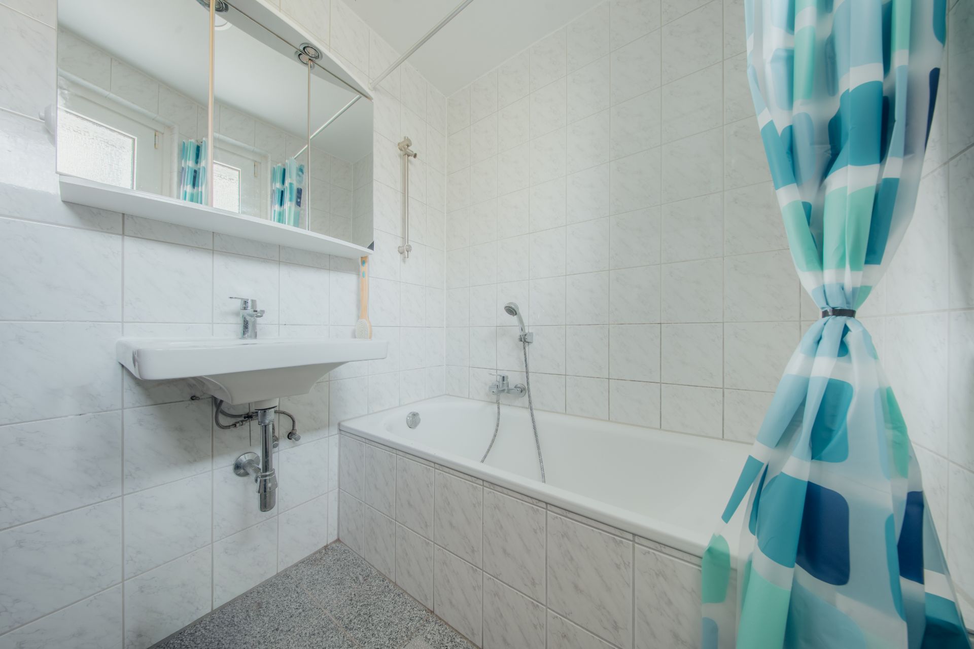 Helles Bad mit Badewanne, Waschbecken, Spiegel und blau gemustertem Duschvorhang.