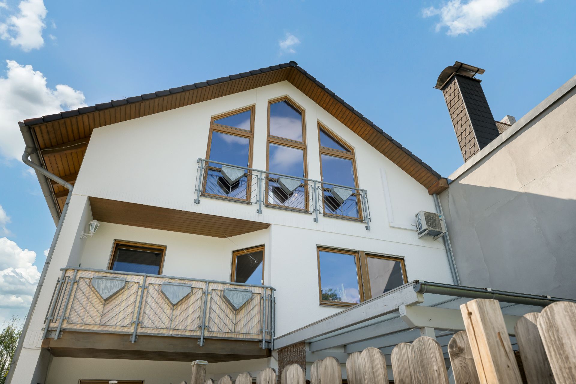 Modernes Haus mit Holz- und Glaselementen, Balkon und markanten Fenstern, davor Holzzaun.