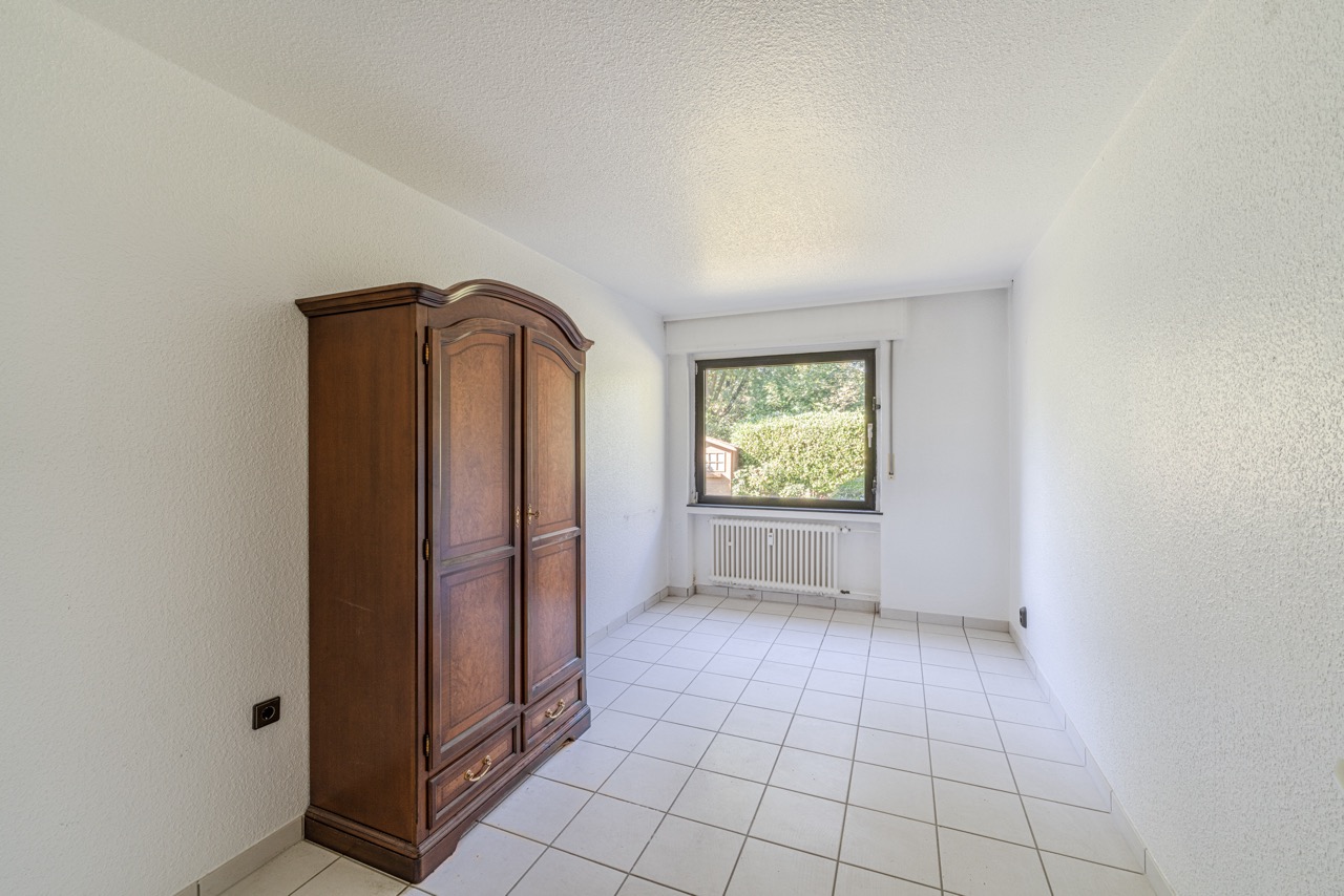 Heller Raum mit Fliesenboden, Holzschrank und Fenster mit Blick ins Grüne.