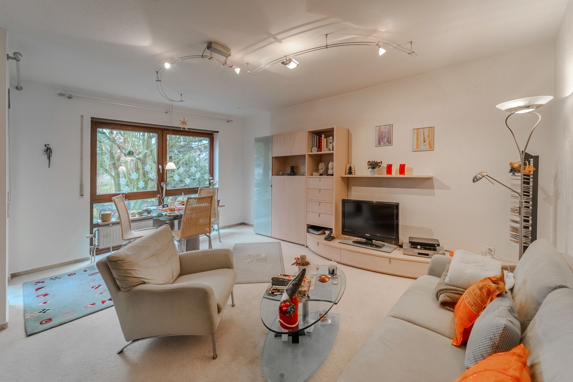 Helles Wohnzimmer mit Sofa, Sessel, Glastisch, TV, Regalwand und Essbereich vor großem Fenster.