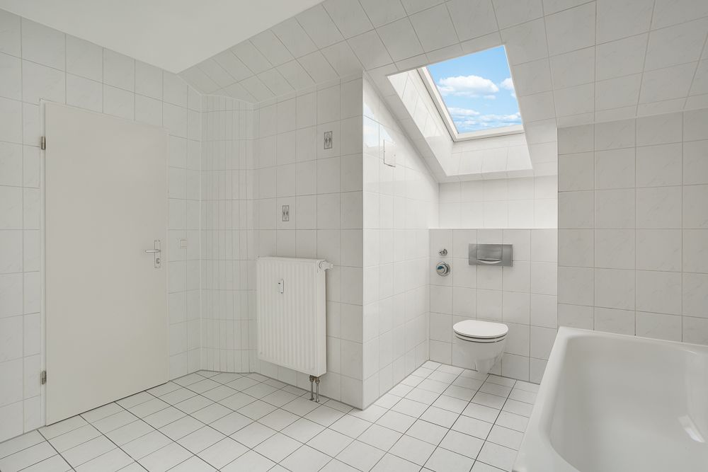 Helles Badezimmer mit weißen Fliesen, Badewanne, WC, Heizung und Dachfenster.