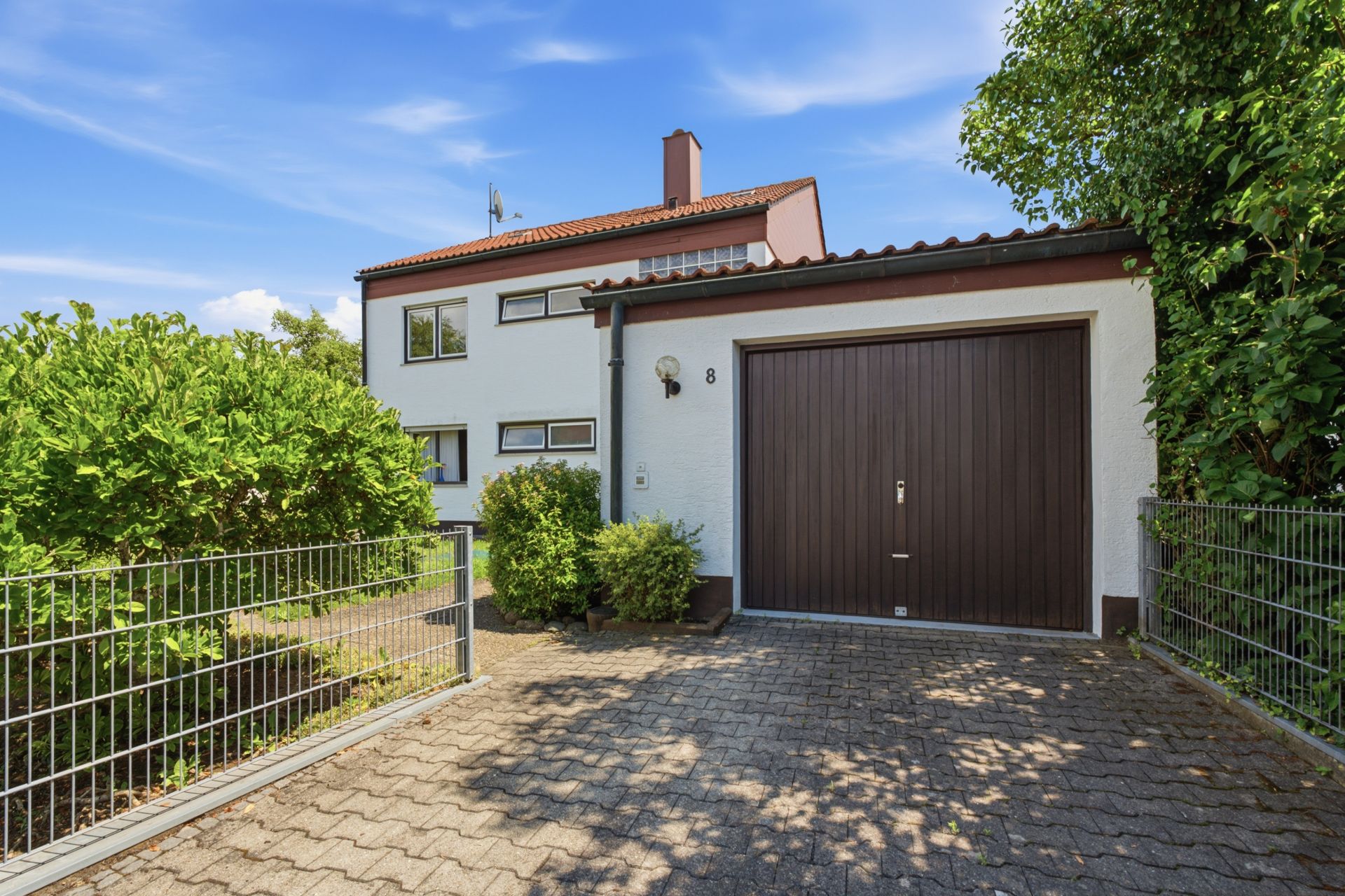 Einfahrt mit Pflastersteinen, Garage und weißes Wohnhaus mit rotem Dach im Hintergrund.