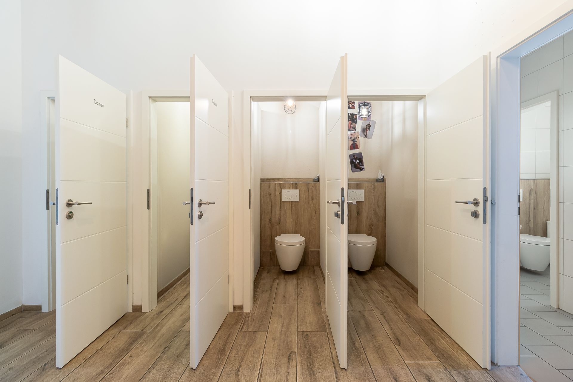 Mehrere offene Türen mit dahinterliegenden Toiletten in einem Flur mit Holzfußboden.