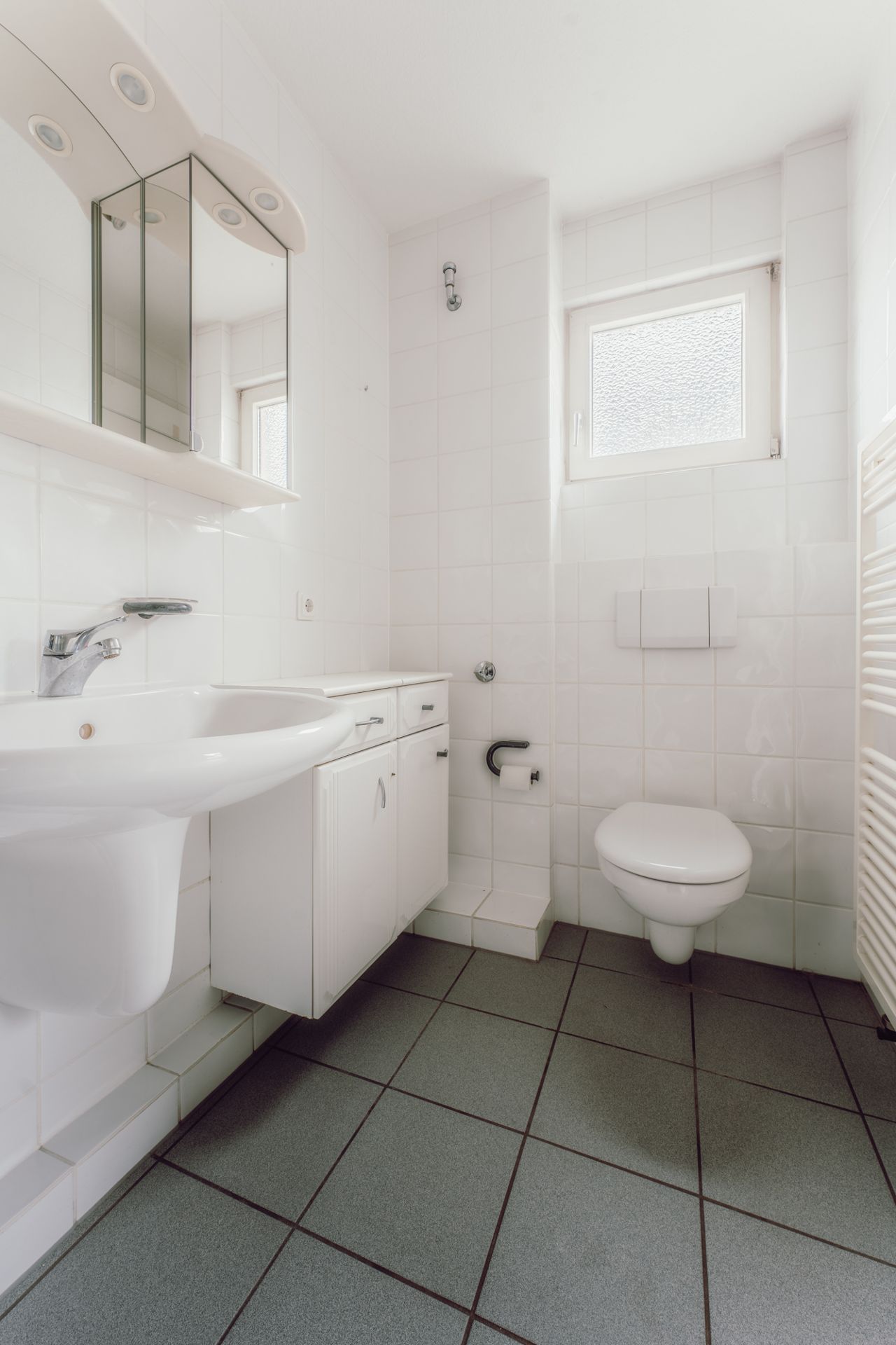 Helles Badezimmer mit Waschbecken, Spiegel, Toilette und Fenster, weiße Fliesen und grauer Boden.
