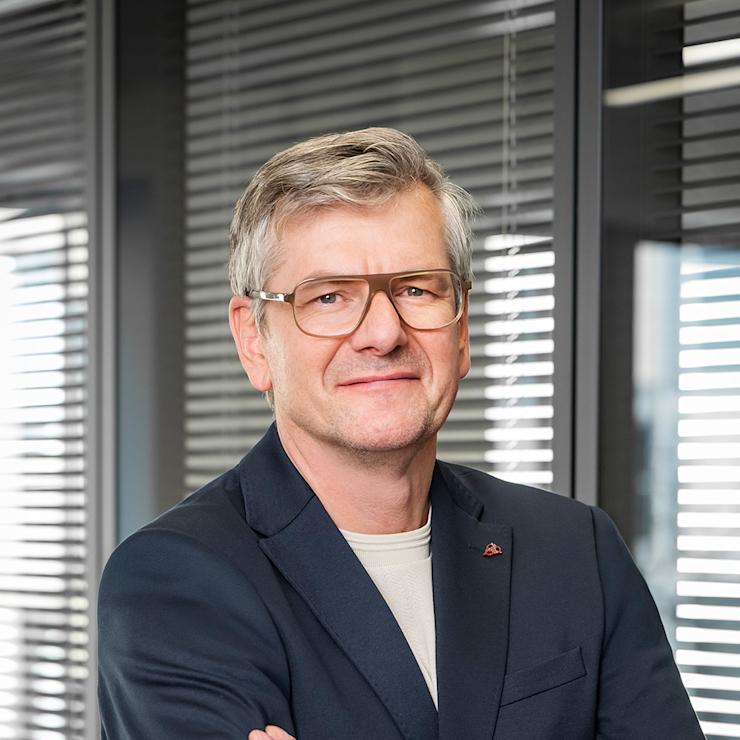 Ein Mann mit Brille und grauem Haar steht mit verschränkten Armen vor Fenstern mit Jalousien.