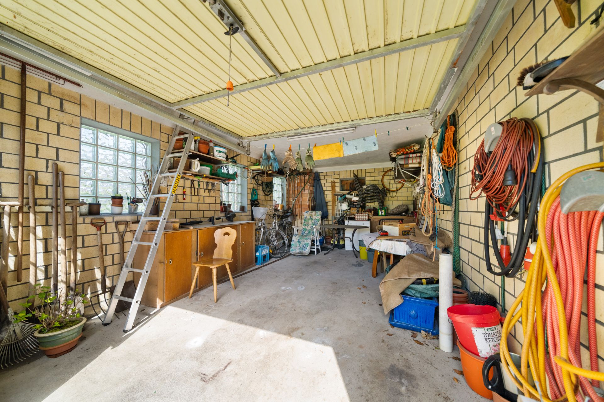 Garage mit Werkzeugen, Leitern, Regalen, Gartengeräten und Fahrrädern an den Wänden.