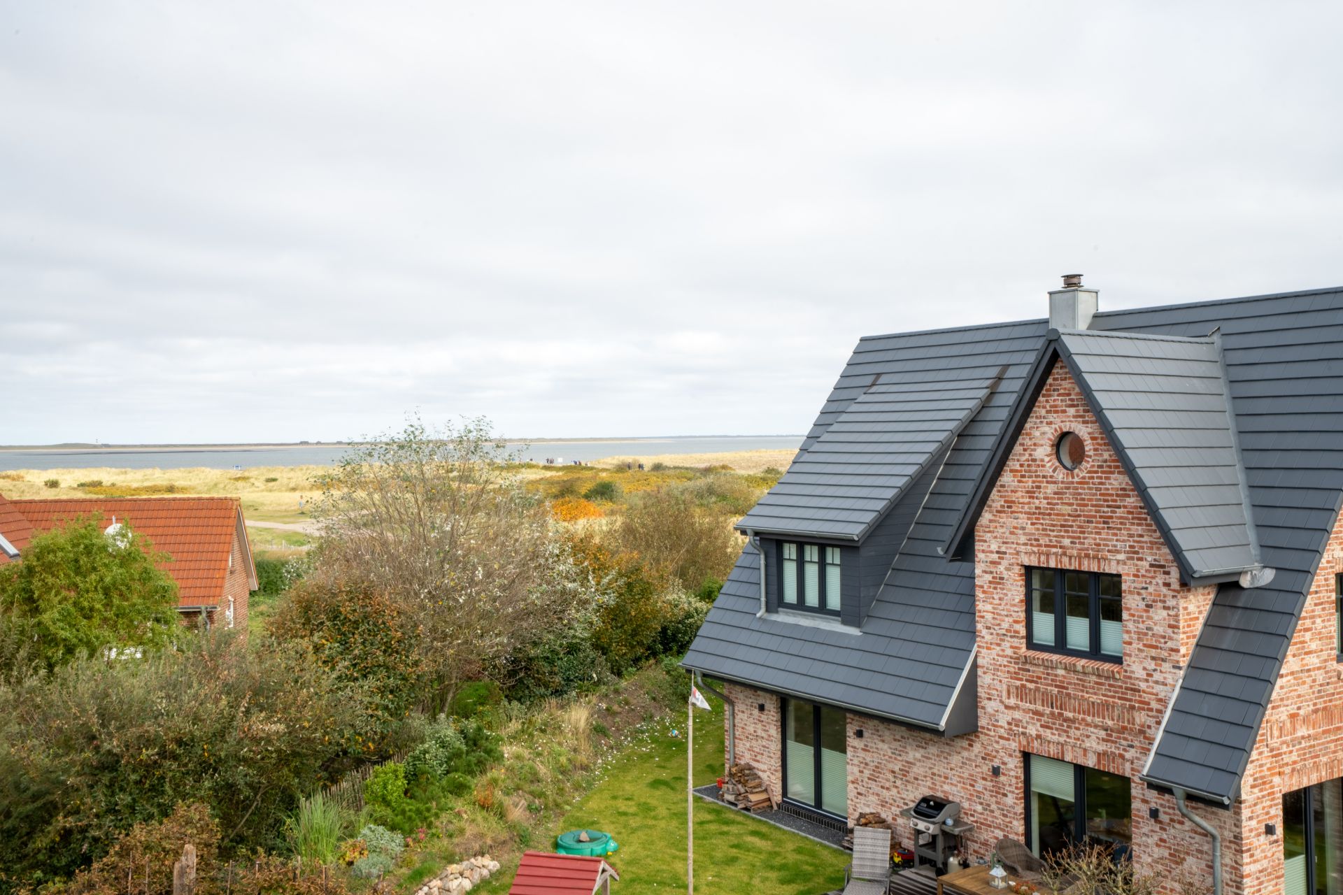 Backsteinwohnhaus mit dunklem Dach, Garten und Blick auf Landschaft mit Wasser im Hintergrund.