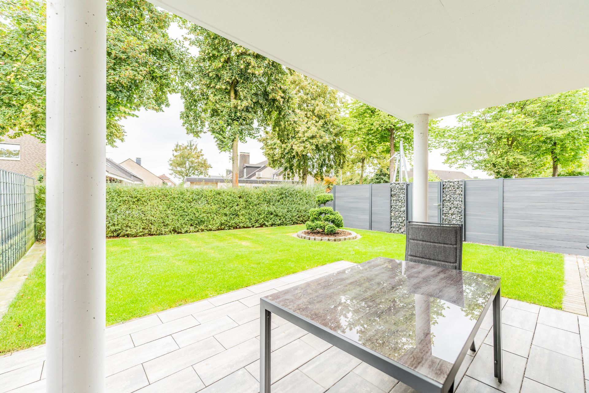 Überdachte Terrasse mit Tisch und Stuhl, Blick auf Garten mit Hecke und Bäumen.
