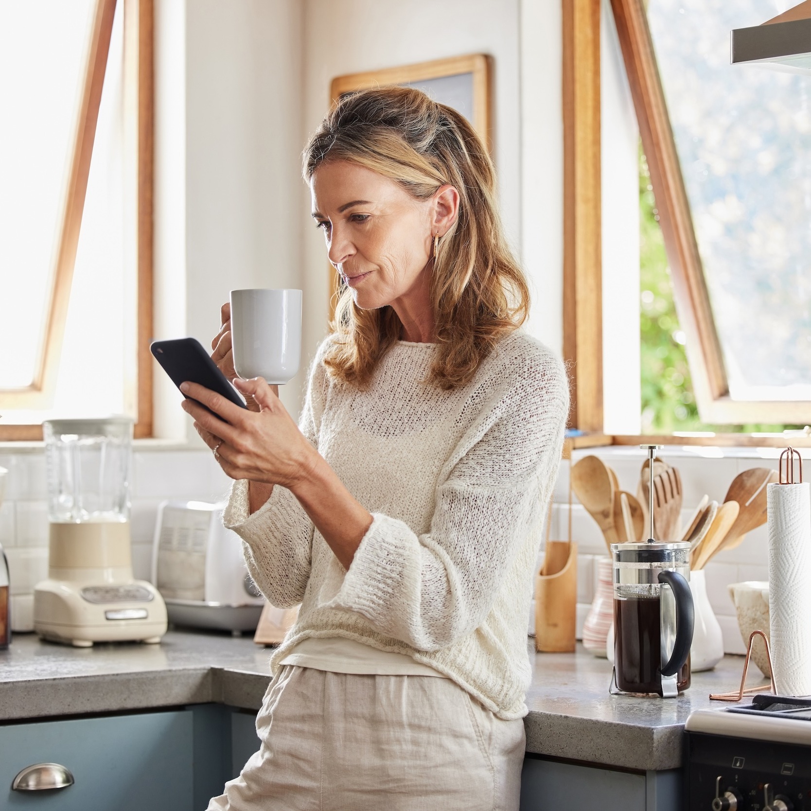 Frau steht in heller Küche, hält eine Tasse und schaut auf ihr Smartphone.