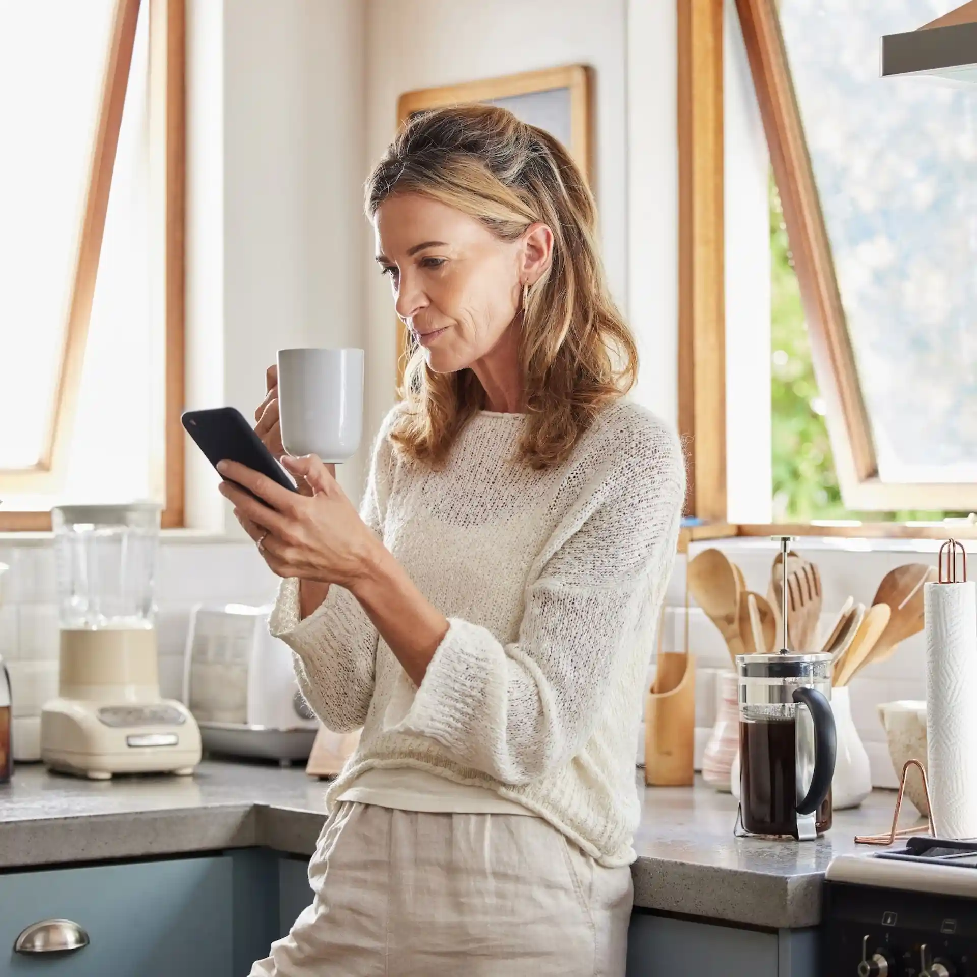 Frau steht in heller Küche, hält eine Tasse und schaut auf ihr Smartphone.