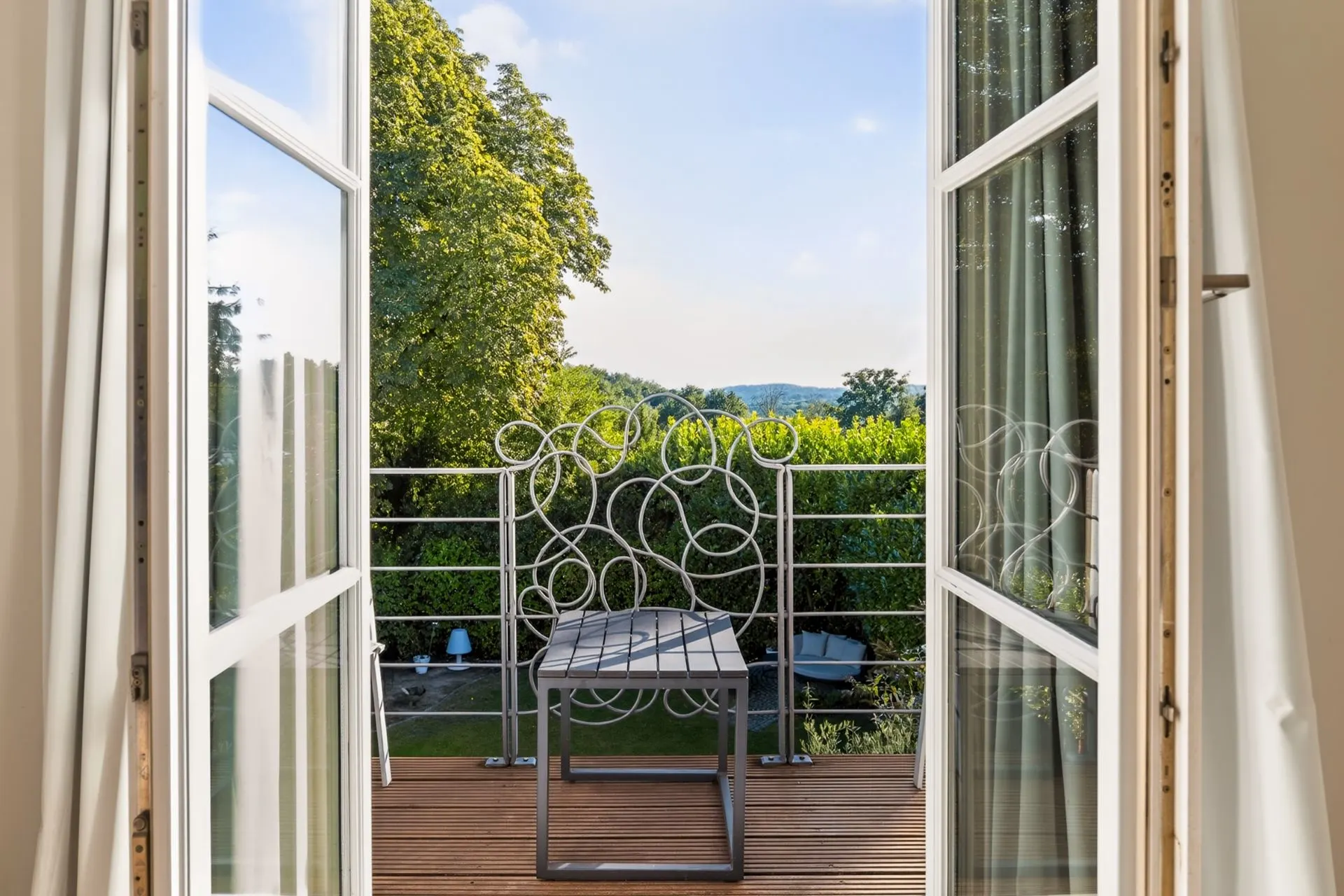 Offene Balkontür mit Blick auf einen kleinen Balkon mit Bank und grünem Garten im Hintergrund.