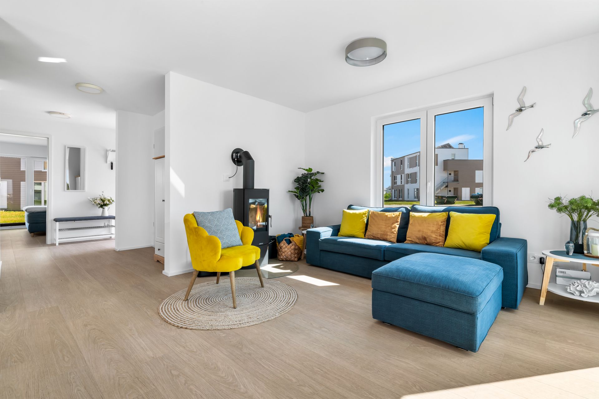 Modernes Wohnzimmer mit blauer Eckcouch, gelbem Sessel, Holzofen und großen Fenstern.