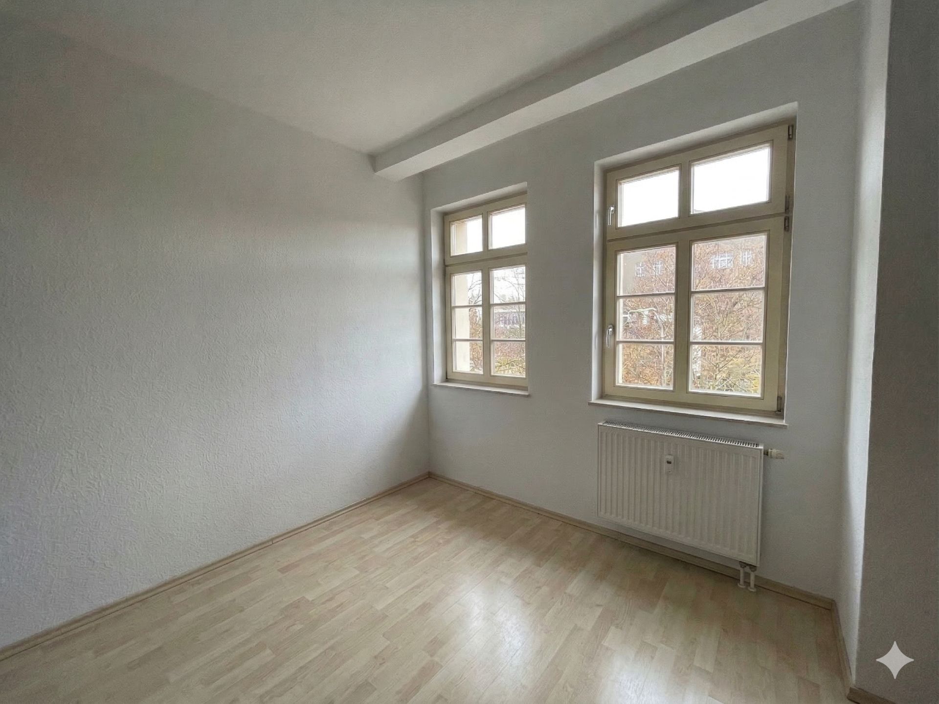 Leerer Raum mit hellen Wänden, zwei Fenstern und Heizkörper auf hellem Laminatboden.