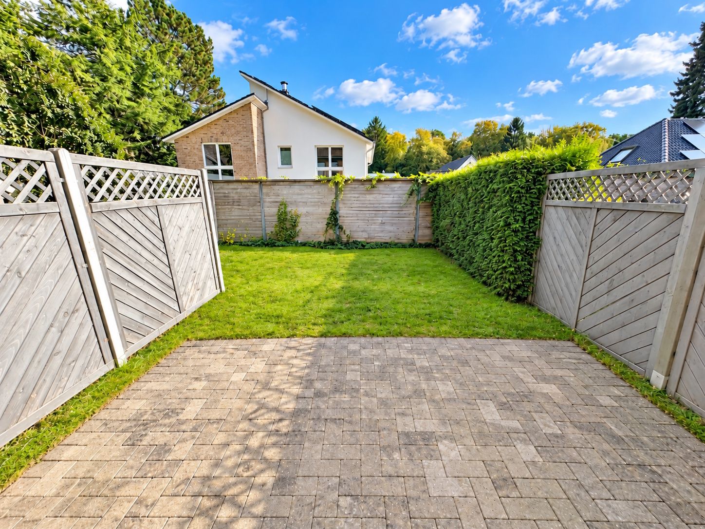 Gepflasterte Terrasse mit Blick auf einen kleinen Garten, umgeben von Holzzäunen und einer Hecke.