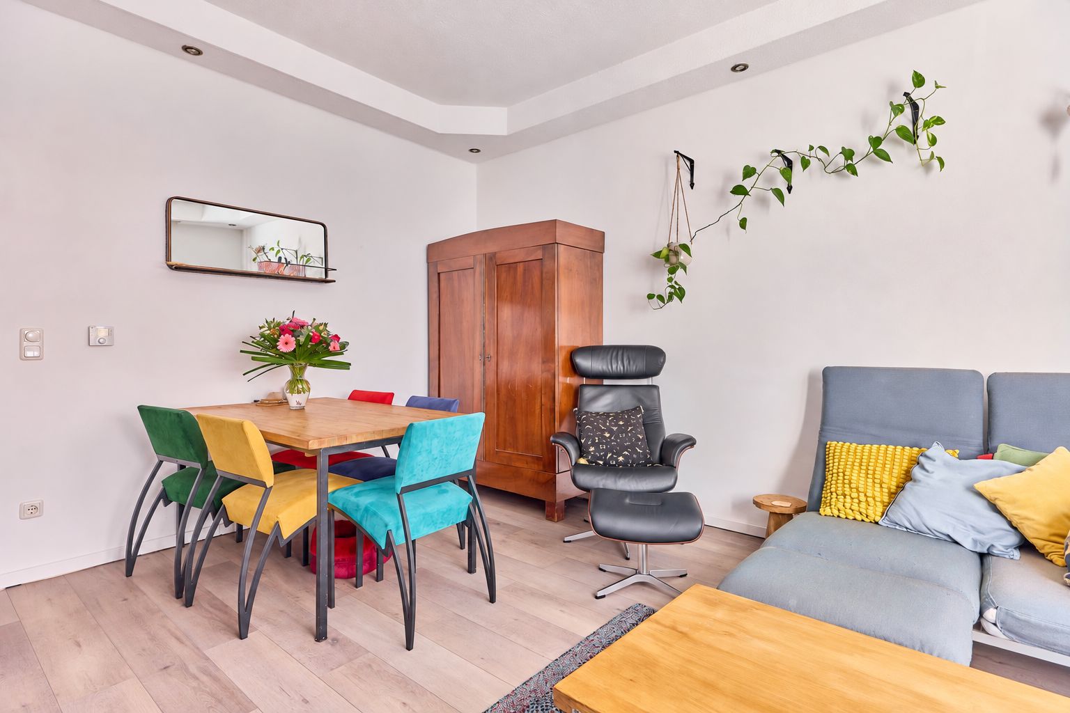Helles Wohnzimmer mit bunten Stühlen, Esstisch, Sofa, Sessel, Holzschrank und Pflanzen an der Wand.