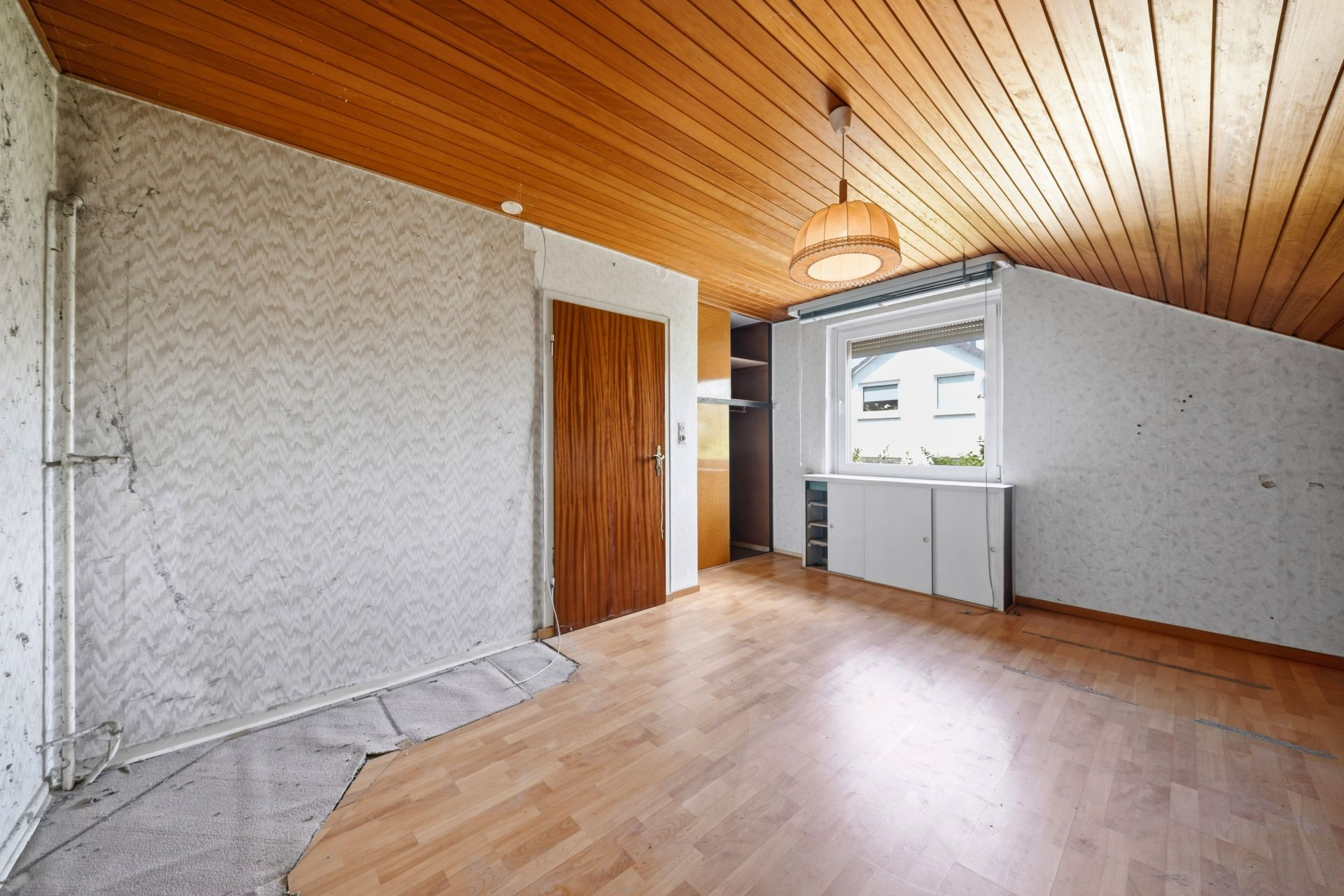 Leeres Zimmer mit Holzdecke, Holzfußboden, kleinem Schrank und Fenster an der rechten Wand.