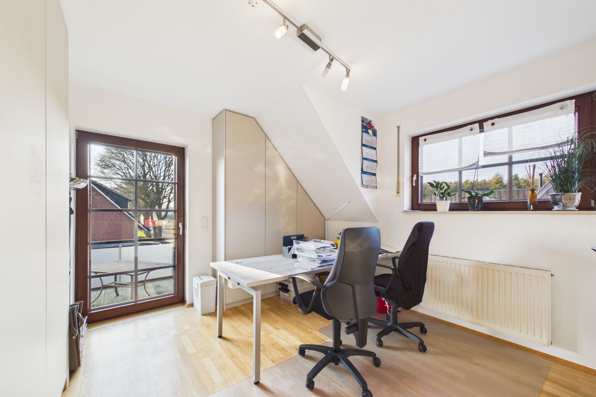 Helles Arbeitszimmer mit zwei Schreibtischen, Stühlen, großen Fenstern und Pflanzen auf der Fensterbank.
