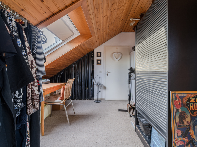 Dachzimmer mit Schrägdecke, Schreibtisch, Stuhl und Kleiderstange, großes Fenster im Dach.