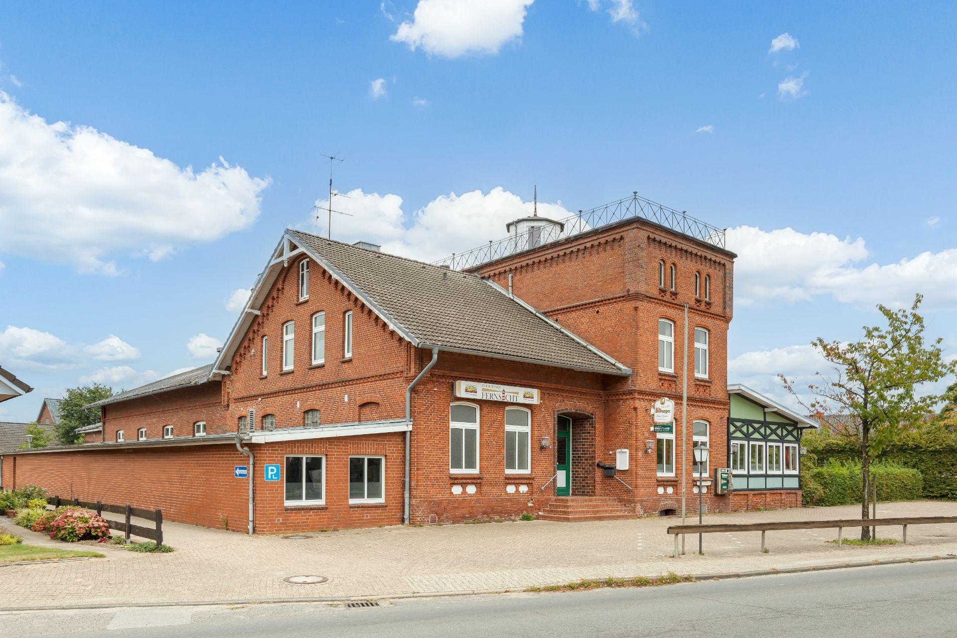 Backsteingebäude mit Satteldach und Turm, davor Gehweg und einige Bäume.