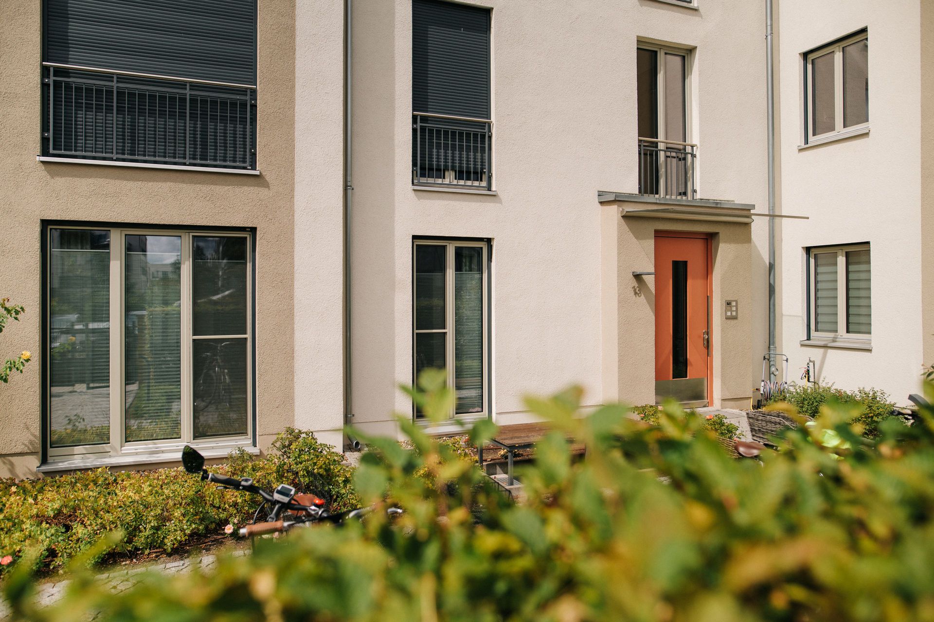 Moderne Wohnhausfassade mit großen Fenstern, Balkon und roter Eingangstür, vorne Sträucher.