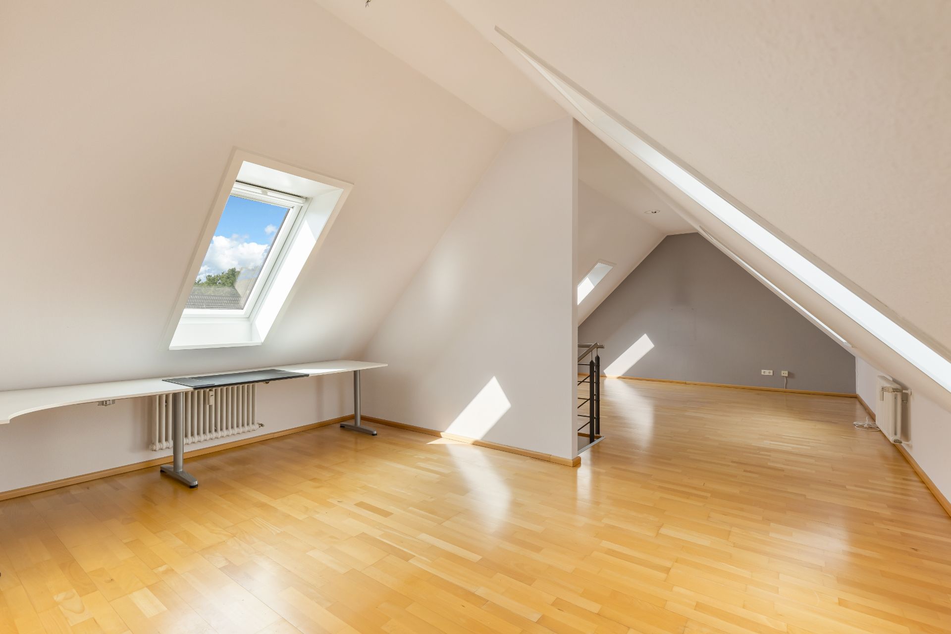 Helles Dachzimmer mit schrägen Wänden, Holzfußboden und mehreren Fenstern.